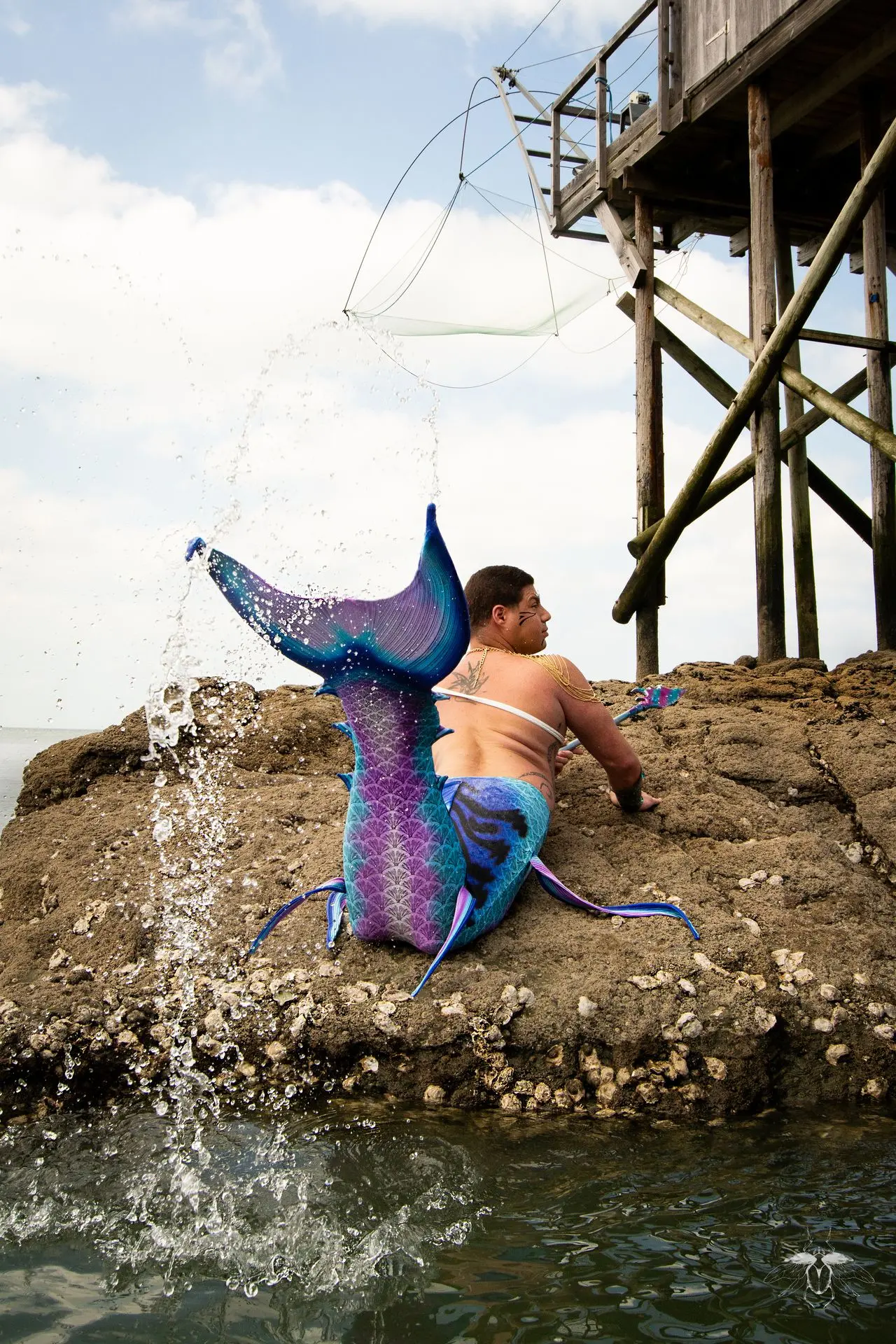 un triton à queue de maquereau rayée, bleu et rose, jaillit hors de l'eau pour se poser sur des rochers avec son harpon.