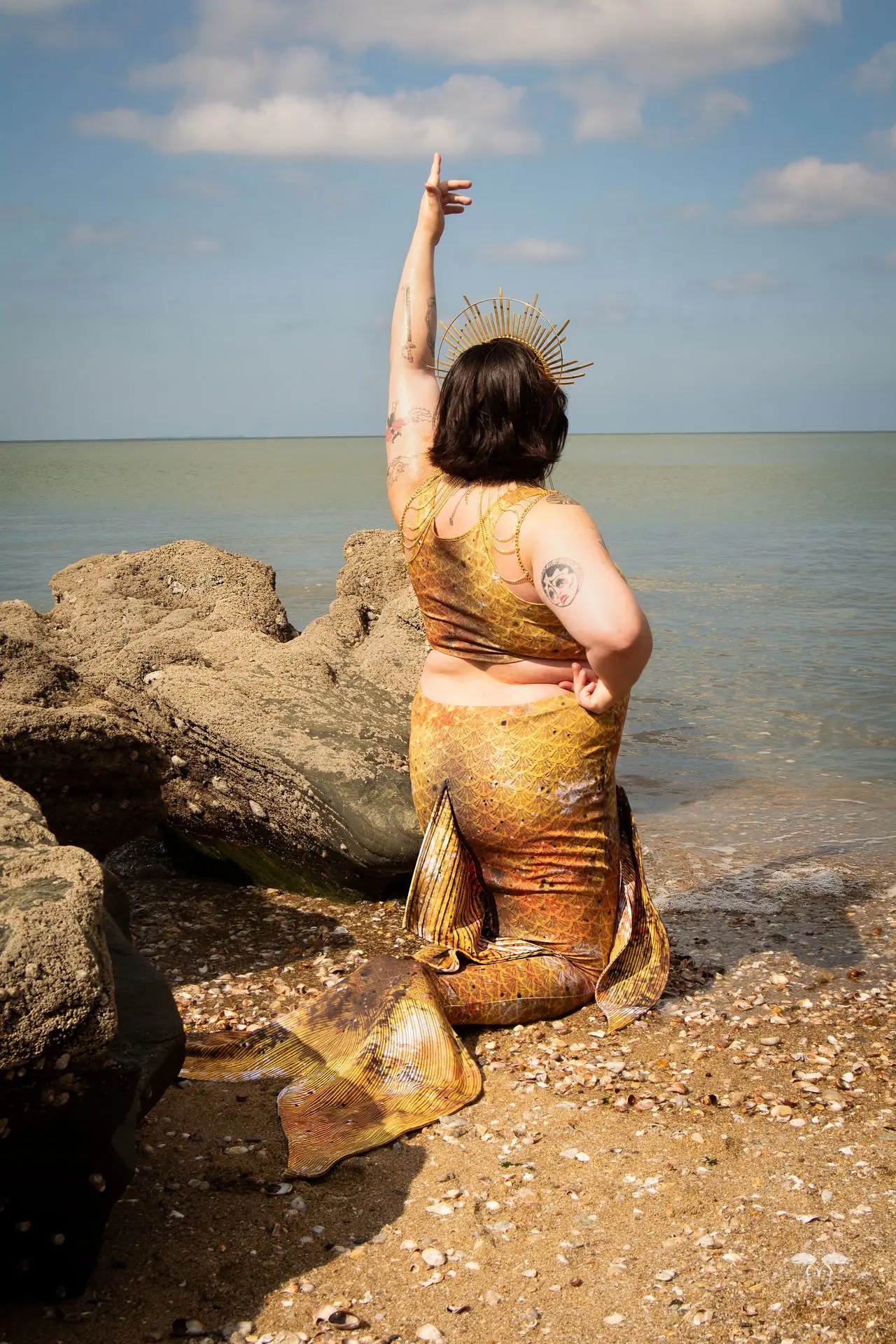 Une sirène jaune et or, couronnée et parée de bijoux dorés, se dresse à genoux vers l'horizon marin, un bras levé pour saluer la beauté de la mer.